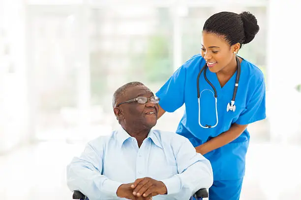 beautiful african nurse taking care of senior patient in wheelchair Premium Care Nurse in blue scrubs with a stethoscope assisting an elderly man in a wheelchair smiling in a bright clinic