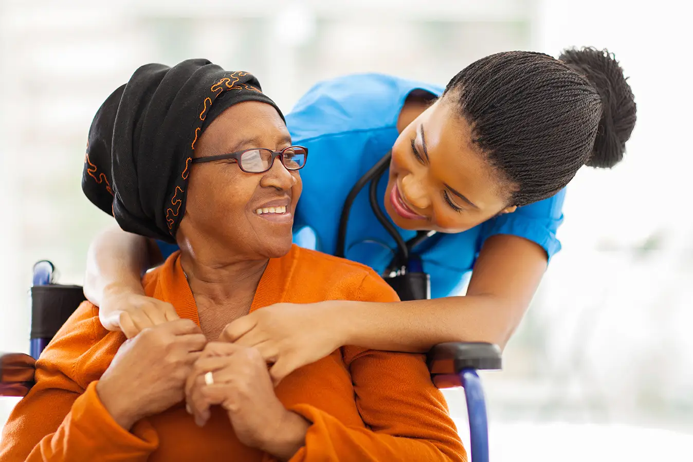 Copy of B2 Premium Care Caregiver nurse in blue scrubs smiles at an elderly woman in a wheelchair sharing a warm moment